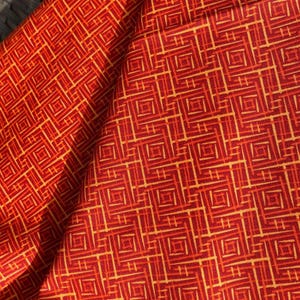 May include: Close-up of a folded fabric with a geometric pattern. The fabric is primarily red with a repeating design of squares and lines in shades of orange and yellow. The fabric appears to be soft and possibly cotton or a similar material.
