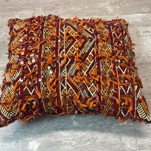 May include: A red, orange, and white patterned throw pillow with fringe and sequins. The pillow is made of a woven fabric and has a geometric design.