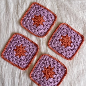 May include: A set of four square crochet coasters featuring a central orange flower motif surrounded by light purple yarn, all bordered by a vibrant orange trim. Each piece is crafted with a textured stitch pattern in a consistent square shape.
