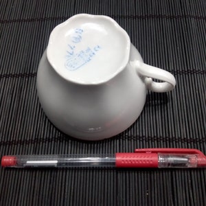 May include: White porcelain teacup with a blue stamp on the bottom that reads "ALL.D.C."