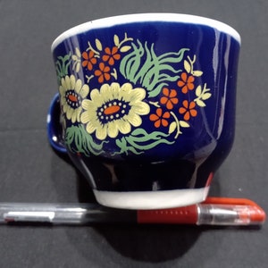 以下が含まれることがあります： 花柄の青いセラミック製のティーカップ。カップは白い縁取りと白い底があります。花は黄色、赤、緑です。