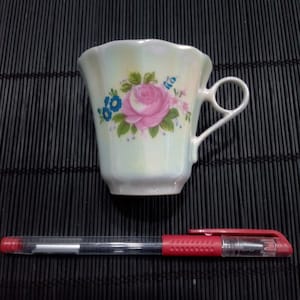 以下が含まれることがあります： ピンクのバラと青い花の模様が入った、真珠色のティーカップ。カップは湾曲したハンドルとスカラップの縁があります。赤と透明のペンがティーカップの下にあります。