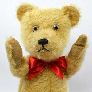 May include: A vintage teddy bear with a red bow tie, wearing a worried expression. The bear has large, black eyes and a brown nose. The bear's fur is a light brown color.