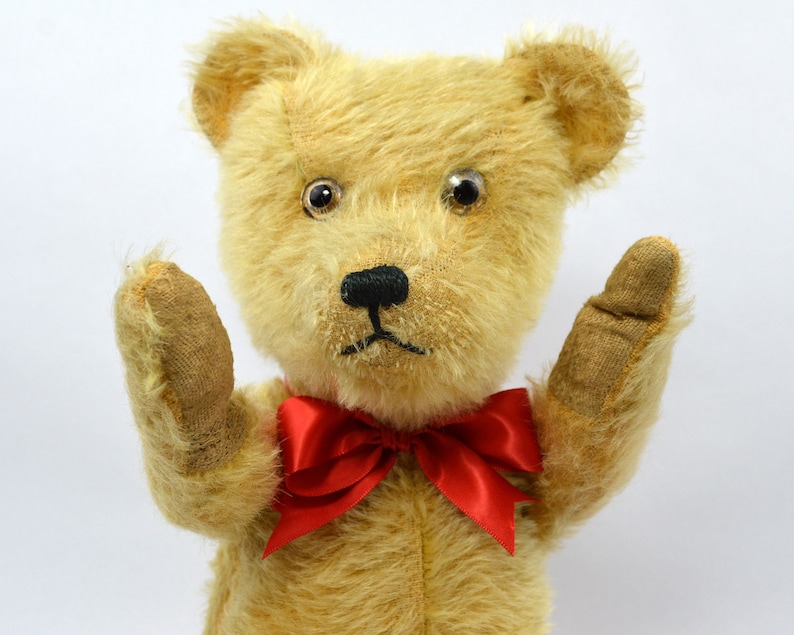 May include: A vintage teddy bear with a red bow tie, wearing a worried expression. The bear has large, black eyes and a brown nose. The bear's fur is a light brown color.