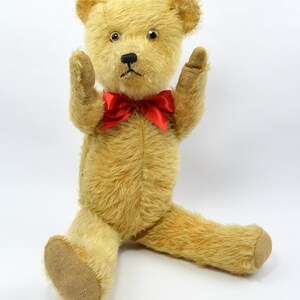 May include: A vintage teddy bear with a red bow tie, sitting with its arms raised. The bear has a light brown fur and a worn, faded appearance.