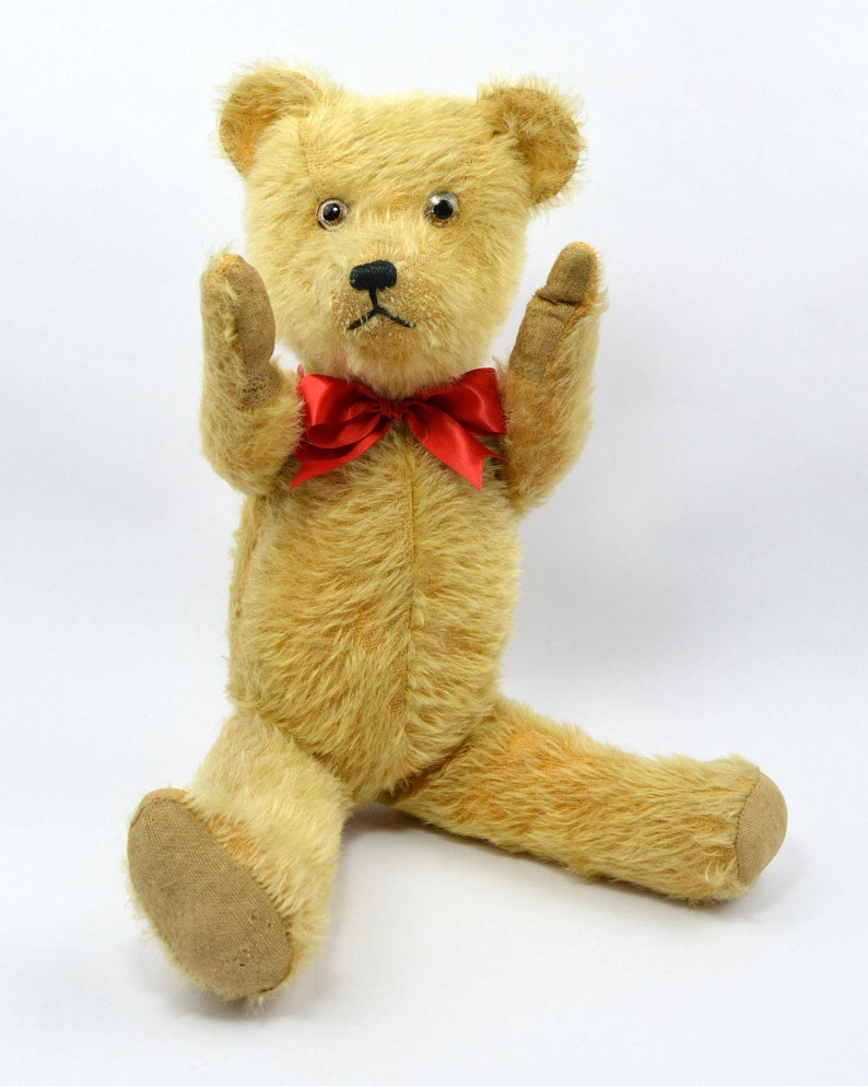 May include: A vintage teddy bear with a red bow tie, sitting with its arms raised. The bear has a light brown fur and a worn, faded appearance.