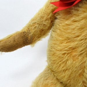 May include: A vintage teddy bear with a worn, tan fur coat and a red bow. The bear's arm is extended, showing the stitching on the paw.