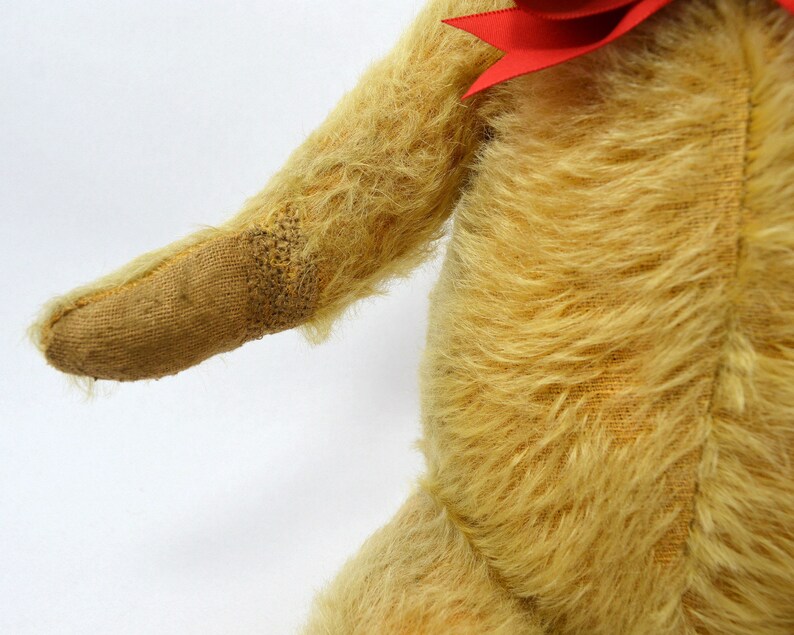 May include: A vintage teddy bear with a worn, tan fur coat and a red bow. The bear's arm is extended, showing the stitching on the paw.
