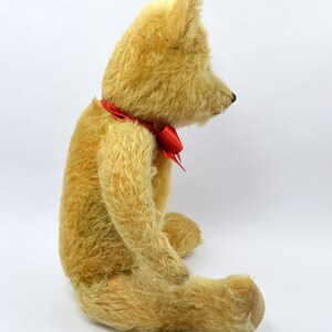 May include: A vintage teddy bear with a cream-colored fur and a red bow tie. The bear is sitting on a white surface.