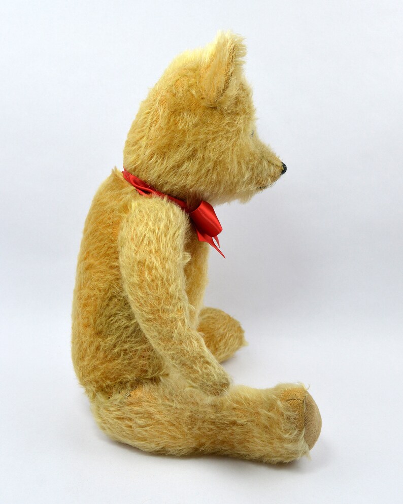 May include: A vintage teddy bear with a cream-colored fur and a red bow tie. The bear is sitting on a white surface.