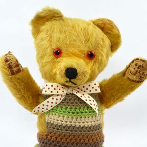 May include: A golden-yellow plush teddy bear with bright red plastic eyes and a black stitched nose. The bear wears a cream-coloured ribbon with small red polka dots tied in a bow around its neck, over a multi-coloured striped crocheted vest.
