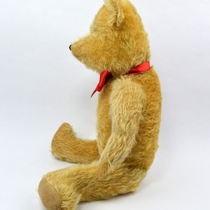 May include: A vintage teddy bear with a light brown fur and a red bow tie. The bear is sitting with its legs crossed and its head tilted to the side.