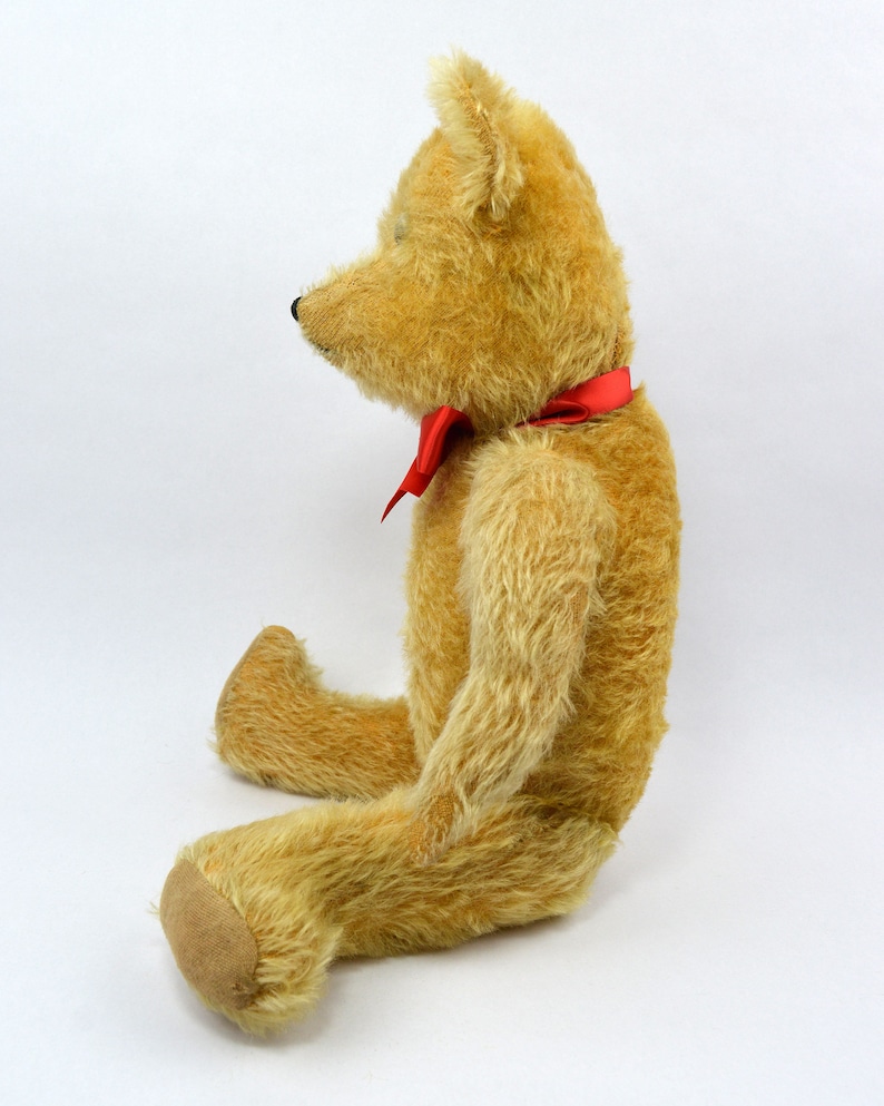 May include: A vintage teddy bear with a light brown fur and a red bow tie. The bear is sitting with its legs crossed and its head tilted to the side.