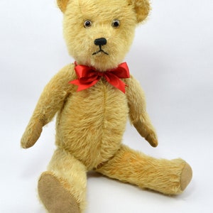 May include: A vintage teddy bear with a light brown fur, black button eyes, and a red bow tie. The bear is sitting with its legs outstretched.
