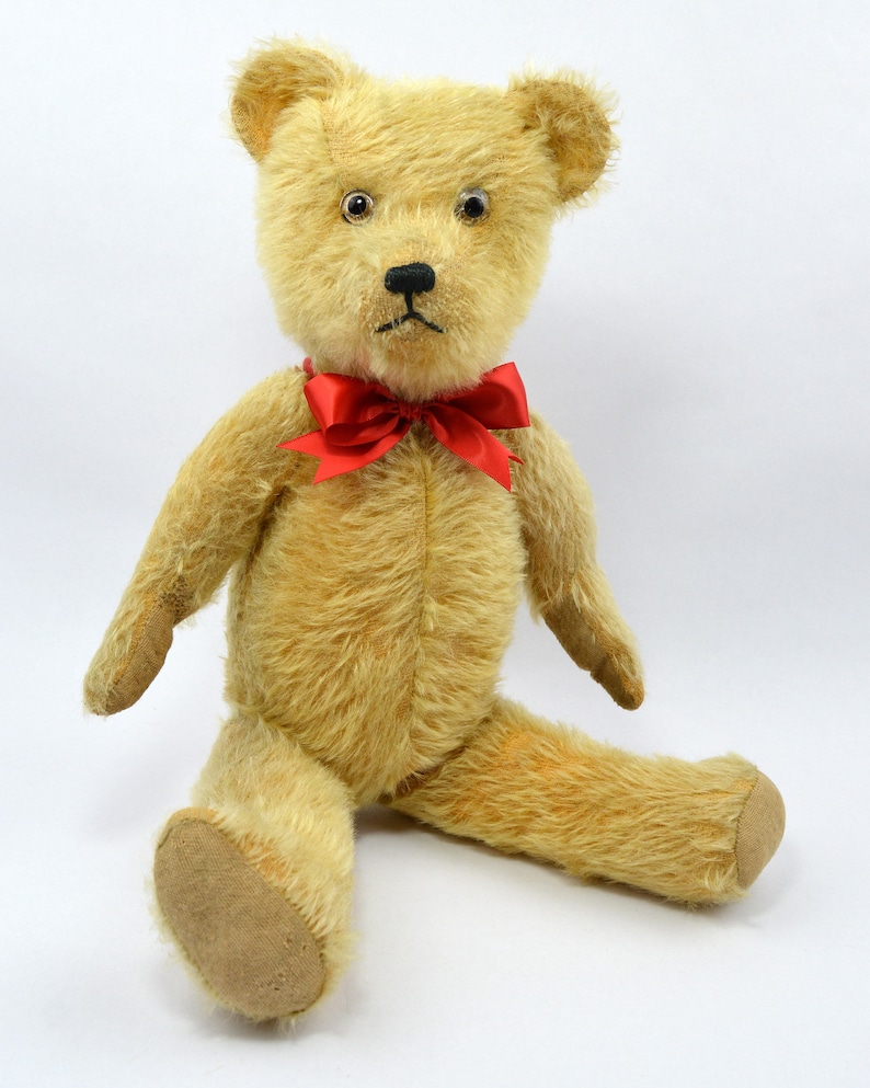 May include: A vintage teddy bear with a light brown fur, black button eyes, and a red bow tie. The bear is sitting with its legs outstretched.