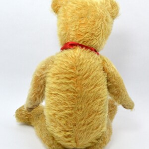 May include: A vintage teddy bear with a light brown fur and a red ribbon around its neck. The bear is sitting on a white background.