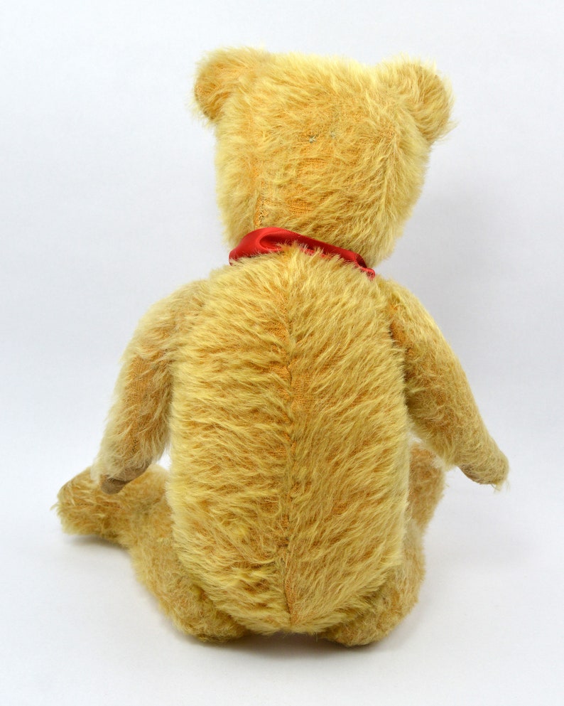 May include: A vintage teddy bear with a light brown fur and a red ribbon around its neck. The bear is sitting on a white background.