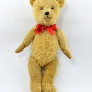 May include: A vintage teddy bear with a light brown fur, black eyes, and a red bow tie. The bear is standing on a white background.