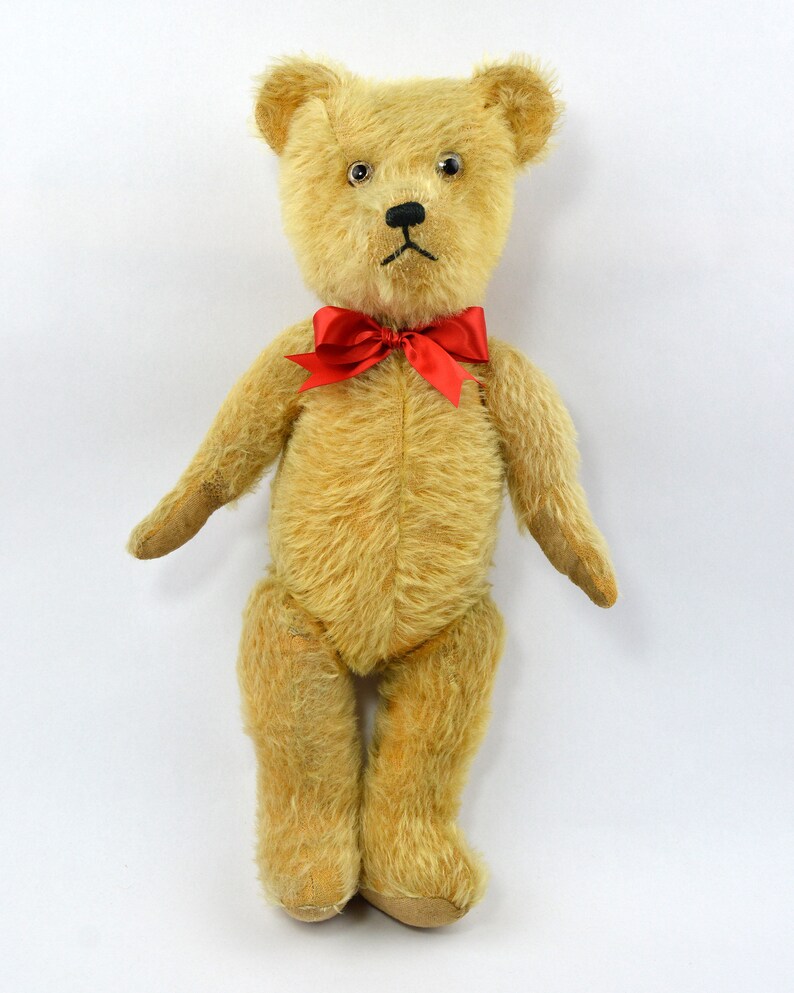 May include: A vintage teddy bear with a light brown fur, black eyes, and a red bow tie. The bear is standing on a white background.