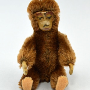 May include: A brown plush monkey toy with a painted face and a long, furry tail. The monkey is sitting with its legs crossed and its arms at its sides.