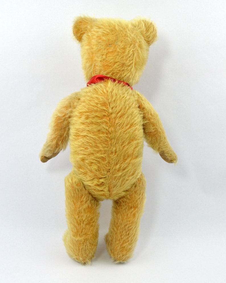 May include: A vintage teddy bear with a red ribbon around its neck. The bear is made of a soft, plush material and has a light brown color.