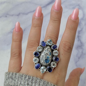 K&ouml;nnte beinhalten: Ein silberner Ring mit einem gro&szlig;en blauen und wei&szlig;en Edelstein, umgeben von kleineren blauen und wei&szlig;en Edelsteinen.