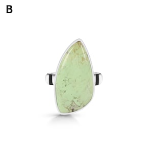 May include: A silver ring featuring a large, teardrop-shaped, light green stone with brown matrix veining. The stone is set in a smooth silver bezel, and the ring band is silver with black accents on the sides. The letter B is visible in the corner.