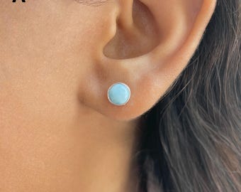 May include: A round stud earring featuring a smooth, light blue stone with subtle white marbling, set in a polished silver-toned metal bezel. The circular gemstone has a flat, polished surface and is secured firmly within the metallic frame.