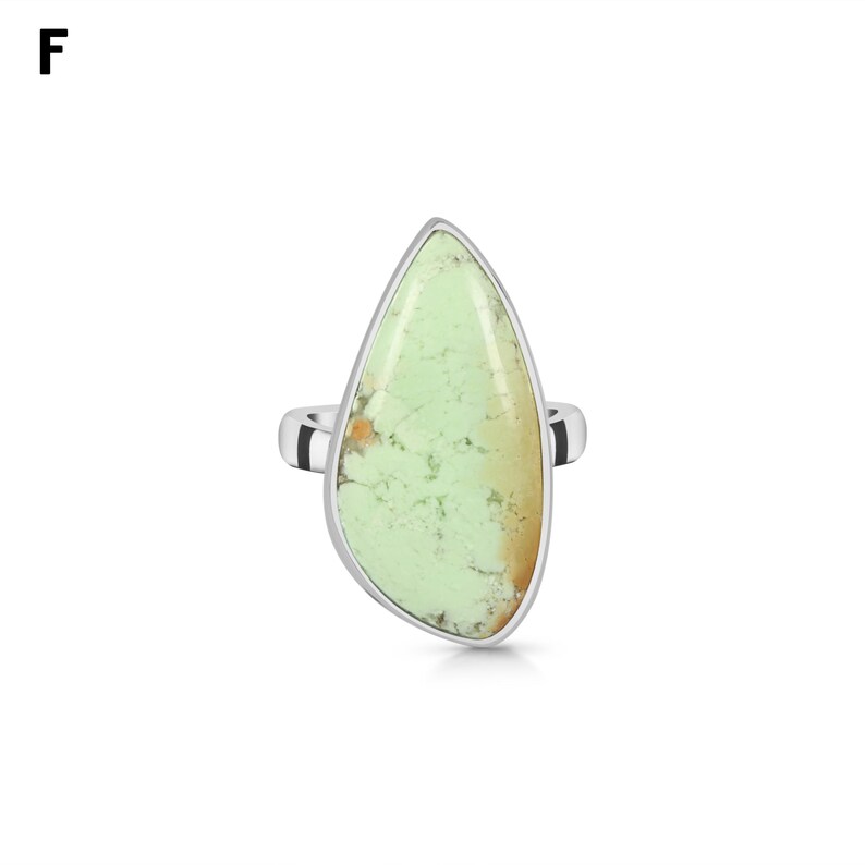 May include: A silver ring featuring a large, teardrop-shaped light green stone with natural dark veining and a small orange-brown inclusion at the base. The stone is set in a smooth, polished silver bezel that follows its irregular shape.
