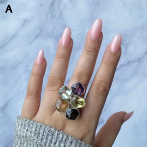 Op de afbeelding: Een zilveren ring met vijf gefacetteerde, veelkleurige edelstenen gerangschikt in een cluster. De stenen hebben tinten paars, lichtgroen, geel, bruin en donkergrijs, allemaal gezet in een gepolijst zilveren metalen frame met letter A.