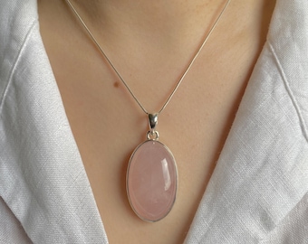 Puede incluir: Un collar con una piedra preciosa de cuarzo rosa pálido, de forma ovalada y lisa, engastada en un bisel de metal plateado pulido. El colgante cuelga de una cadena fina y delicada de color plateado que descansa sobre la zona de la clavícula.