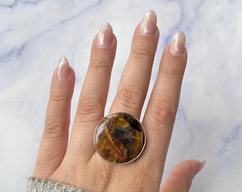 May include: A circular ring featuring a polished, multi-colored stone with swirling patterns of brown, gold, and dark blue. The stone is set within a simple, thin silver-toned metal band that encircles the circumference of the round gemstone.