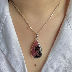 Può includere: Una collana d'argento con un pendente a goccia. Il pendente presenta una pietra preziosa rosa e nera, incastonata in una lunetta d'argento. La collana è mostrata su una camicia bianca.