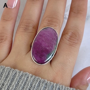 Puede incluir: Un anillo ovalado grande con una piedra pulida de color magenta intenso con inclusiones naturales oscuras y un sutil tono verde en la base, engastado en una banda de metal plateado simple y lisa que rodea toda la piedra preciosa.