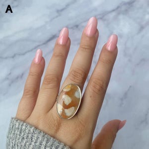 May include: A silver ring featuring a large, oval-shaped cabochon stone with a mottled pattern of warm orange, cream, and white tones. The stone is set in a smooth, polished silver bezel, creating a bold statement piece that sits prominently on the finger.