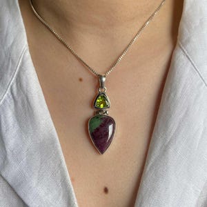 May include: A silver necklace with a pendant featuring a teardrop-shaped green and pink gemstone, accented with a smaller triangular green gemstone.