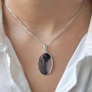 May include: A silver chain necklace with an oval pendant featuring a dendritic agate stone. The stone has a black and white pattern resembling a tree.