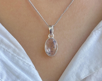 Op de afbeelding: Een zilveren ketting met een ovaalvormige hanger van een heldere, doorschijnende steen met kleine donkere insluitingen. De hanger zit vast aan een eenvoudig zilveren oogje en hangt aan een fijne zilveren ketting tegen een witte achtergrond.