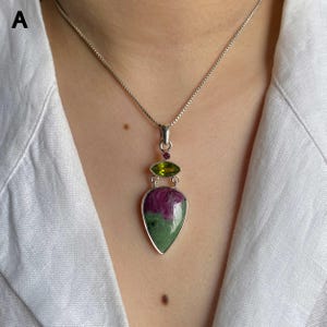 May include: A silver pendant necklace featuring a large, teardrop-shaped ruby zoisite stone with green and magenta hues. Above the main stone sits a smaller, marquise-cut lime green peridot and a tiny round pink gemstone, all set in silver.