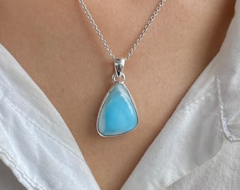 May include: A teardrop-shaped blue Larimar gemstone pendant set in a polished silver frame, suspended from a delicate silver chain. The stone features a smooth, opaque surface with swirling patterns of light and sky blue hues against a white shirt.