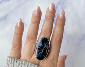 May include: A silver ring featuring a large, oval-shaped stone with a dark blue, black, and gold marbled pattern. The stone is set in a smooth, polished silver bezel, creating a bold statement piece that covers a significant portion of the finger.