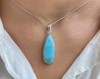 May include: A teardrop-shaped light blue Larimar gemstone pendant set in a polished silver frame, suspended from a thin silver snake chain necklace. The stone features natural swirling patterns in shades of sky blue and white, with a smooth finish.