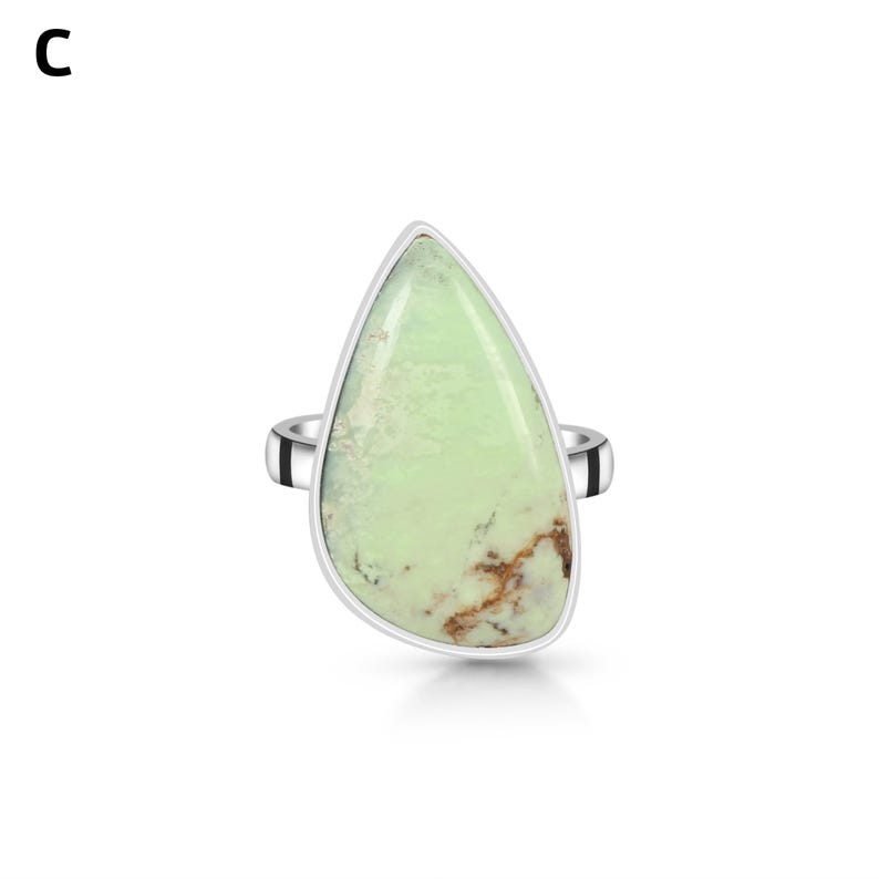 May include: A silver ring featuring a large, teardrop-shaped, light green stone with brown matrix inclusions. The stone is set in a smooth silver bezel, and the band is a simple, polished silver metal. The letter C is visible in the top left corner.