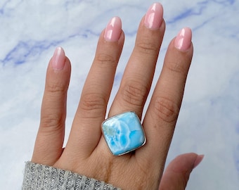 May include: A square-shaped ring featuring a polished, light blue and white marbled stone set in a silver-toned metal frame. The stone displays a natural, cloud-like pattern with varying shades of sky blue and white, secured by a smooth, metallic bezel.