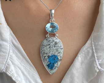 Könnte beinhalten: Eine silberne Anhängerkette mit einem tropfenförmigen Stein, der eine gesprenkelte grau-weiße granitartige Textur und einen leuchtend blauen Einschluss aufweist. Darüber sitzt ein kleinerer, oval geschliffener hellblauer Edelstein in Silber.