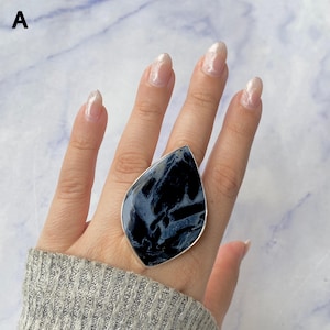 May include: A large teardrop-shaped ring featuring a dark blue and black marbled stone set in a polished silver-toned metal frame. The stone displays intricate, swirling patterns of deep navy and charcoal grey throughout its smooth surface.