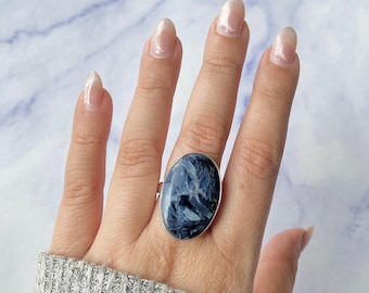 May include: A ring featuring a large, oval-shaped blue and grey stone with a marbled, chatoyant pattern set in a simple silver-toned metal band. The stone displays deep navy, slate blue, and white streaks throughout its polished, reflective surface.