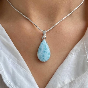 Puede incluir: Un collar de plata con un colgante en forma de lágrima. El colgante presenta una piedra preciosa azul claro con vetas blancas. El collar se muestra sobre una camisa blanca.