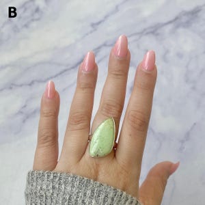 May include: A ring featuring a large, teardrop-shaped, light green stone with dark veining set in a simple metal band. The stone has a smooth, polished surface and is positioned on the ring finger, showcasing its distinct colour and natural pattern.