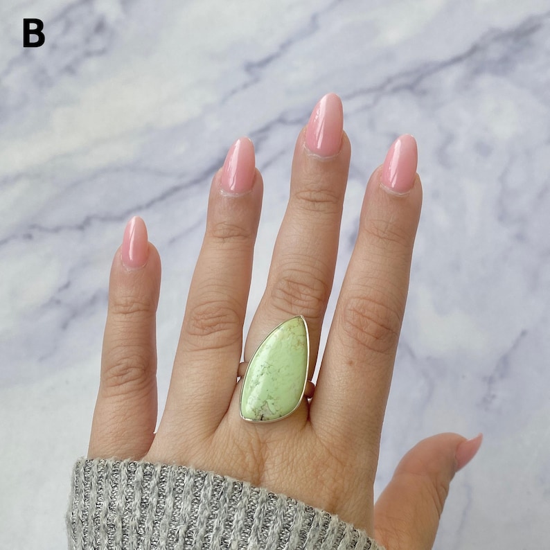 May include: A ring featuring a large, teardrop-shaped, light green stone with dark veining set in a simple metal band. The stone has a smooth, polished surface and is positioned on the ring finger, showcasing its distinct colour and natural pattern.